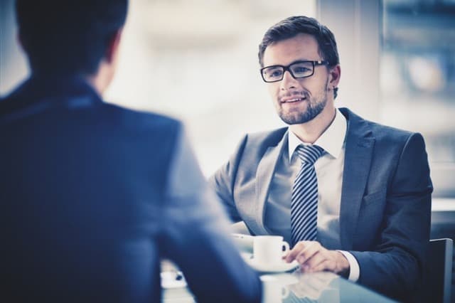 Homem sentado em uma mesa com uma xícara de café conversando com outro homem